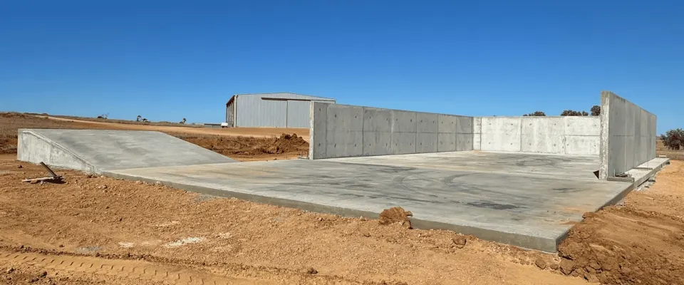 A finished concrete slab with tilt-panel walls and an access ramp on red dirt, with a farm shed behind