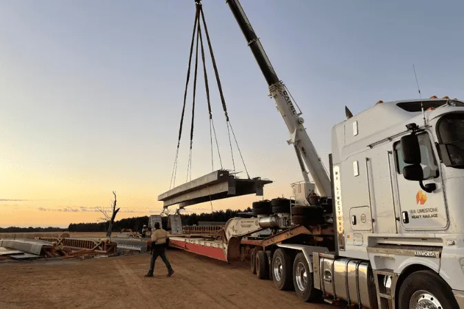 A crane lifting a precast bridge beam off a truck at sunset